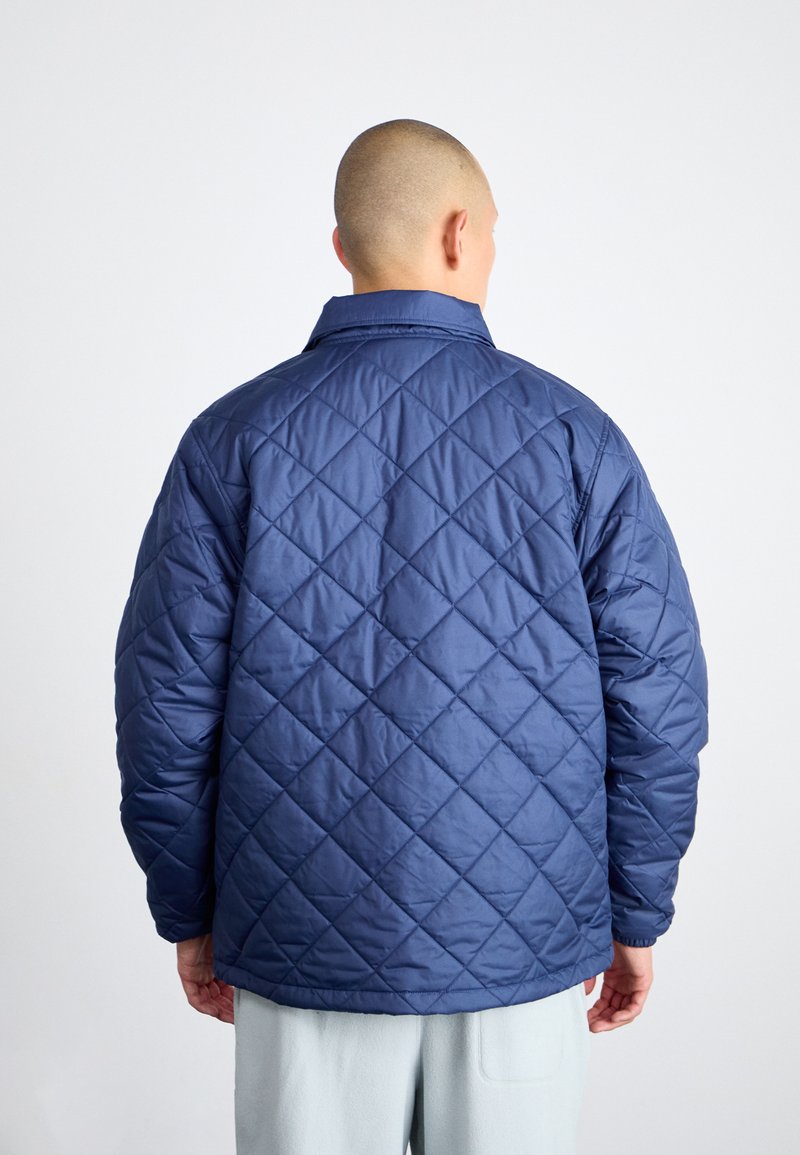 Veste matelassée bleu marine avec un motif en losange, un col standard et des manches à coupe décontractée. Tissu lisse et léger sans matériel visible.