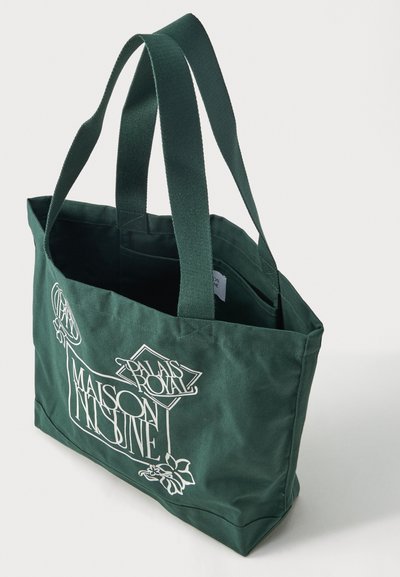 Mørkegrønn lerretveske med to håndtak og hvit tekstdesign med teksten "Palais Royal Maison Insigne" med dekorative elementer.