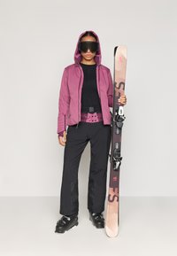 Ski oblečení zahrnující vínovou bundu, černý základní oděv, černé kalhoty a lyžařské boty, držící vzorované lyže s vázáním.