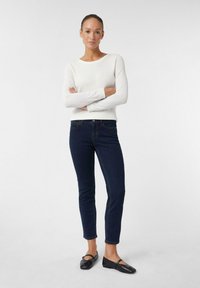 Witte, textuurgevoelige longsleeve trui met een ronde hals, gedragen met strakke donkerblauwe jeans en zwarte platte schoenen. Staand met de armen gekruist.