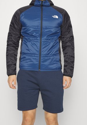 Blauwe en zwarte gewatteerde jas met ritssluiting, capuchon en het logo van The North Face op de borst, gedragen met marineblauwe korte broek.