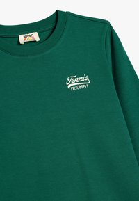 Grøn sweatshirt lavet af bomuld. Har en rund halsudskæring og et broderet "Tennis Triumph" logo i hvidt. Glat tekstur, afslappet design.