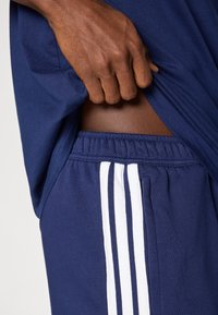 Αθλητικό σορτς σκούρου μπλε με λευκές κάθετες ρίγες στα πλάγια· απαλό ύφασμα· ε λαστική μέση· χέρι που κρατά το πουκάμισο στη μέση.