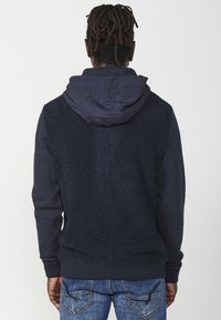 Hoodie med en mjuk, texturerad marinblå baksida och en slät, marinframsida. Inkluderar en huva med dragsko och ribbade ärmslut.