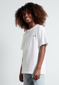 Tricou alb din bumbac cu mâneci scurte, având un mic logo verde pe partea stângă a pieptului și o croială lejeră. Purtați cu blugi albaștri deschiși.