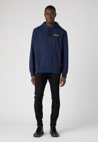 Sudadera con capucha de color azul marino, mezcla de algodón, con bolsillo frontal, capucha ajustable mediante cordones, logo bordado en amarillo, combinada con pantalones negros de corte slim y botas.