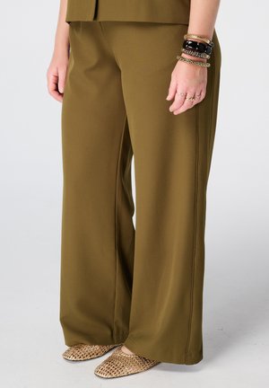 Femme portant un pantalon large olive, des chaussures plates tissées dorées et plusieurs bracelets à son poignet droit avec des bagues à ses doigts.