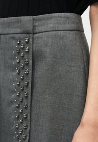 Γκρι φούστα με κομψό σχεδιασμό, διακοσμημένη με ασημένιες καρφίτσες κατά μήκος της μιας πλευράς. Το ύφασμα έχει λεπτή υφή, που ενισχύει τη συνολική της εμφάνιση.