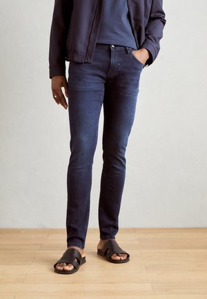 Person iført mørke skinny jeans, mørke slidssandaler og en mørk jakke, der står på lyst trægulv mod en ensfarvet væg.