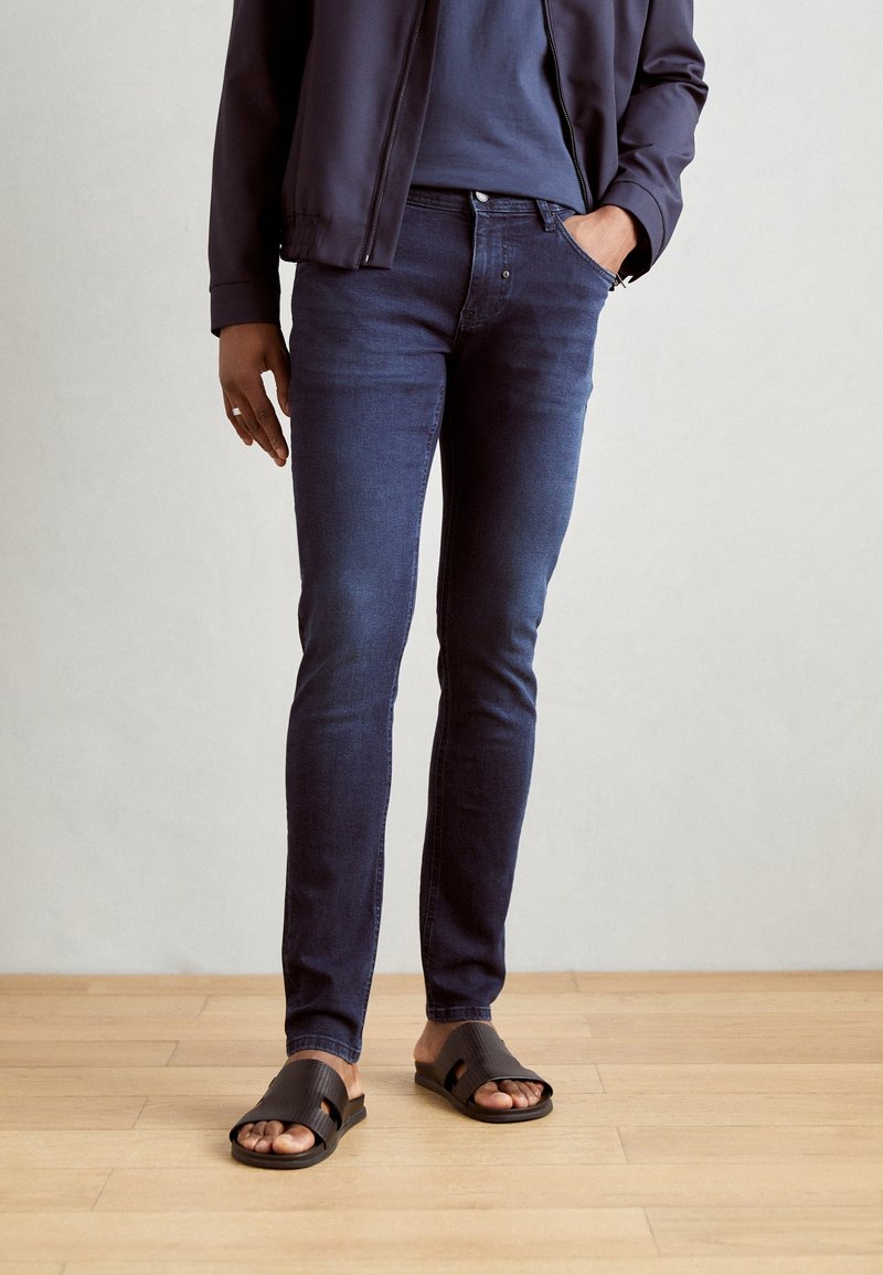 Personne portant un jean skinny foncé, des sandales slides foncées et une veste foncée, debout sur un sol en bois clair contre un mur uni.