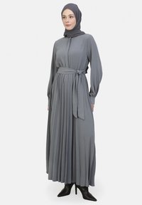 Langes, plissiertes Kleid aus weichem grauem Stoff, mit einem gebundenen Taillengürtel, Knopfleiste und langen Ärmeln, kombiniert mit schwarzen High-Heels.