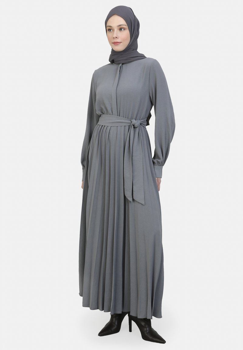 Langes, plissiertes Kleid aus weichem grauem Stoff, mit einem gebundenen Taillengürtel, Knopfleiste und langen Ärmeln, kombiniert mit schwarzen High-Heels.