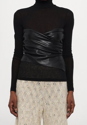 Femme portant un col roulé noir transparent à manches longues, un haut enveloppant en cuir noir et un pantalon en tricot crème.