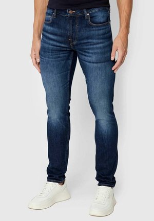 Homme portant un jean slim bleu foncé et des baskets blanches, debout devant un fond clair uni.