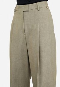 Pantaloni beige chiaro in tessuto testurizzato. Presentano una vita alta, pince frontali e una tasca laterale, rifiniti con un piccolo accento metallico.