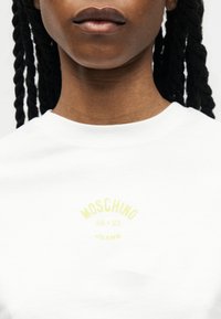 Κοντινό πλάνο ατόμου που φοράει λευκό t-shirt Moschino Jeans με ανοιχτό κίτρινο λογότυπο, εμφανίζοντας τον λαιμό, τα χείλη και τα πλεγμένα μαλλιά.