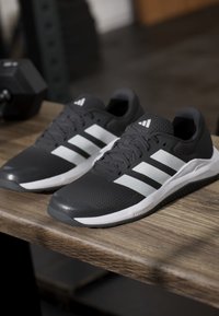 Chaussures de course Adidas noires et blanches avec des bandes blanches, posées sur une surface en bois à côté d'un haltère noir dans une salle de sport.