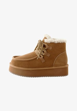 Tan mockasiner ankelboots med tjock sula, fuskpälsfoder och dekorativa snören framtill; rundad tå och strukturdetaljer.