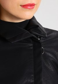 Chaqueta de cuero negra con un cuello estructurado, que presenta un acento de metal y una textura visible. Apariencia suave y elegante.
