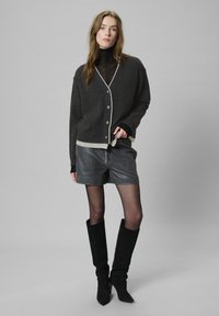 Cárdigan de punto gris con ribetes en crema contrastantes, diseño abotonado, combinado con pantalones cortos de cuero gris y botas altas de ante negras.