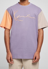 Fialové tričko s krátkým rukávem, které má oranžové a béžové rukávy, s texturovaným vyšitým logem v kurzívě přes prsa.