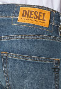 Jean en denim avec une étiquette en cuir beige marquée "DIESEL" à l'arrière à la taille, avec des coutures jaunes et une texture légèrement usée.
