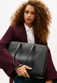 Γυναίκα με σγουρά μαλλιά που φοράει μπορντό σακάκι και λευκό πουκάμισο, κρατώντας μια μεγάλη μαύρη τσάντα με υφή, με χρυσό λογότυπο και λαβές.