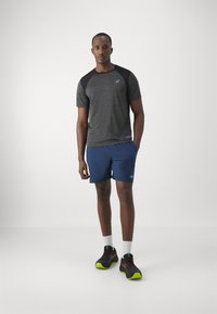 Gray short-sleeve athletic shirt with black shoulder accents, paired with blue shorts and black shoes with neon green soles. White socks visible.