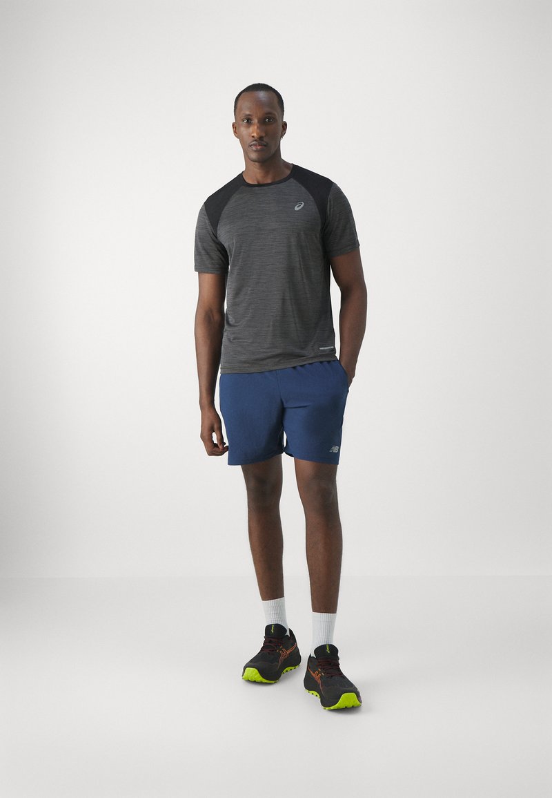 Camiseta atlética gris de manga corta con acentos negros en los hombros, combinada con pantalones cortos azules y zapatos negros con suelas de color verde neón. Calcetines blancos visibles.