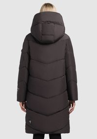 Person som bär en lång svart pufferjacka med en stor huva, sedd bakifrån mot en enkel ljus bakgrund.