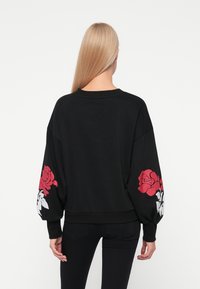 Sudadera negra de manga larga con gráficos de rosas rojas y blancas en las mangas. Material suave, corte relajado, dobladillo acanalado.
