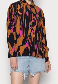 Langærmet skjorte i sort med abstrakte orange og pink mønstre. Har en knapkrave og mansjettdetaljer. Kombineret med en denim nederdel.