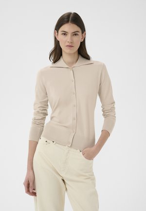 Jonge vrouw met steil bruin haar draagt een beige vest met knopen en crème-kleurige broek, staand met één hand in de zak tegen een effen achtergrond.