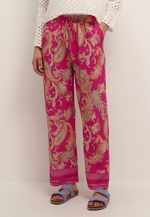 Mujer con pantalones anchos de color rosa brillante con patrones intrincados de cachemira, un suéter de punto blanco y sandalias azules.