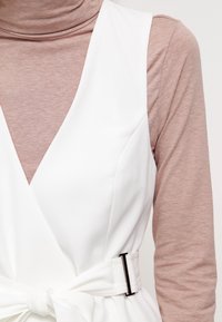 Biała sukienka bez rękawów z głębokim dekoltem w kształcie litery V, wykonana z teksturowanej tkaniny, z paskiem w talii i czarną metalową klamrą. Nosi się ją na jasnobrązowym golfie.