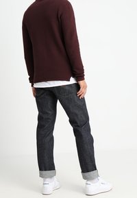 Bordeauxroter strukturierter Pullover, dunkle Jeans mit hochgekrempelten Bündchen und weiße Sneakers, von hinten gesehen, wobei der Farbkontrast des Outfits hervorgehoben wird.
