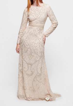 Mujer que lleva un vestido largo de noche beige con cuentas, con patrones intrincados y mangas largas, posando con una mano en la cadera.