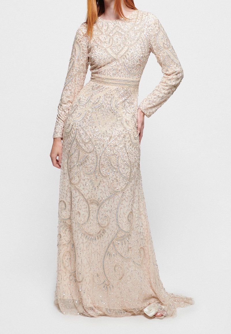 Femme portant une longue robe de soirée beige ornée de perles avec des motifs complexes et des manches longues, debout avec une main sur la hanche.