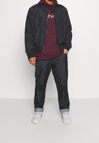 Chaqueta bomber negra sobre un hoodie burdeos, combinada con jeans de mezclilla oscuros enrollados en los puños y zapatillas blancas. Diseño simple, sin patrones.