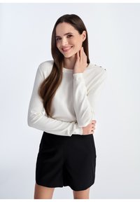 Kremowy sweter z długim rękawem o okrągłym dekolcie i złotymi guzikami na ramionach, w połączeniu z czarną spódnicą. Gładka faktura.