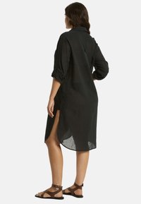 Una donna con capelli ondulati, vestita con un abito nero a maniche lunghe e sandali marroni, sta in piedi con le spalle alla fotocamera, mostrando delle aperture laterali.