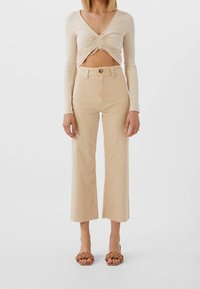 Haut beige à manches longues avec un devant torsadé, associé à un pantalon beige taille haute à jambes larges avec un ourlet effiloché, porté avec des sandales marron à semelles épaisses.