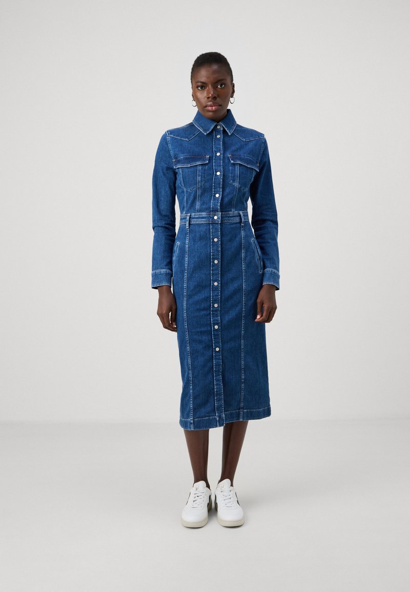 Denimklänning i mörkblått, långa ärmar, knapprad fram, två bröstfickor, bälte i midjan, knälång, matchad med vita sneakers.