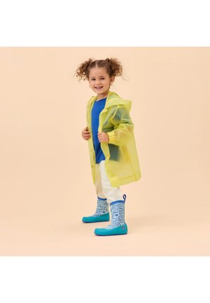 Junges Mädchen lächelnd, trägt einen gelben Regenmantel, blaues Hemd, weiße Hose und blaue, gemusterte Gummistiefel, steht vor einem beigen Hintergrund.