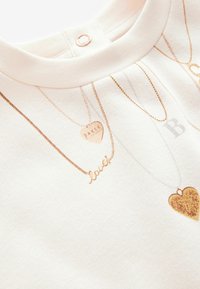 Weißes T-Shirt aus Stoff mit goldfarbener Druckgestaltung von Halsketten, einschließlich eines Herzens mit "BAKER", einer "love" Schrift und einem glitzernden Herzakzent.