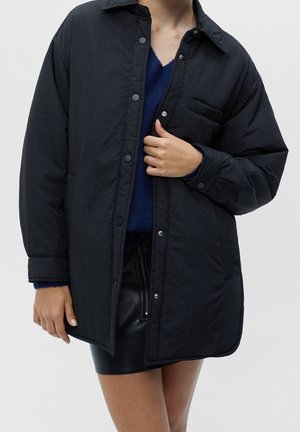 Person trägt eine schwarze gesteppte Jacke mit Knöpfen über einem blauen Pullover und einem schwarzen Lederrock, Hand richtet die Jacke nahe der Brusttasche.