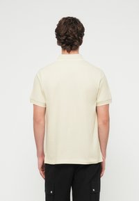 Άνδρας με κατσαρά μαλλιά, ντυμένος με λευκό, απλό κοντομάνικο πόλο και μαύρο παντελόνι cargo, εμφάνιση από πίσω με φόντο λευκό.