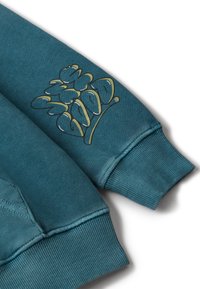 Tyrkysová mikina s elastickým manžetovým rukávem, na kterém je stylizovaný zlatý a černý graffiti potisk na látce.