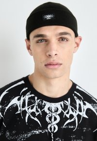 Black knit headband with a small logo. Model wears a black t-shirt featuring a white graphic design with abstract shapes and textures.