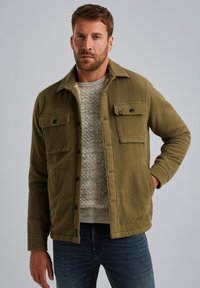 Mężczyzna w oliwkowej kurtce zapinanej na guziki, noszący jasnoszary sweter z dzianiny i ciemnoniebieskie dżinsy, stoi z jedną ręką w kieszeni.